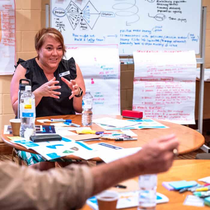 Changemaker facilitator, Tanya Lehman sits at a table of participants in a well lit room. Behind her is a whiteboard with ideas generated from the workshop.