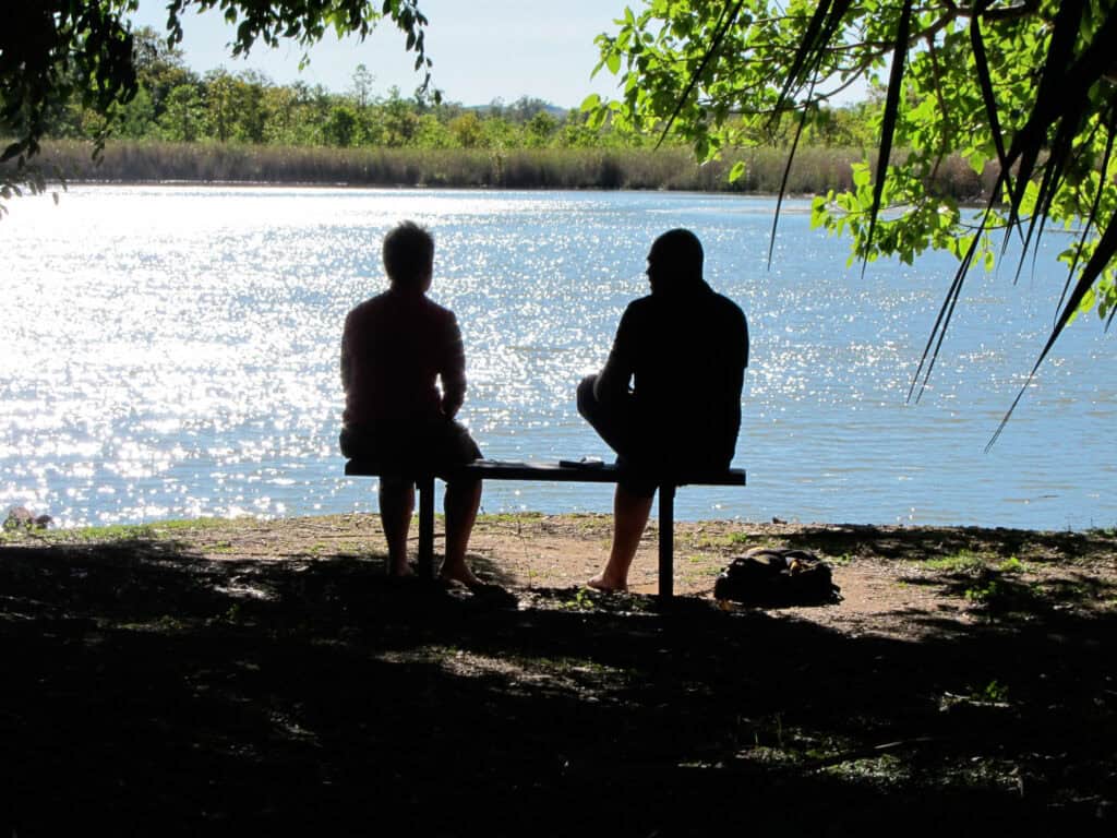 Two men sitting in the shadows in front of a river.