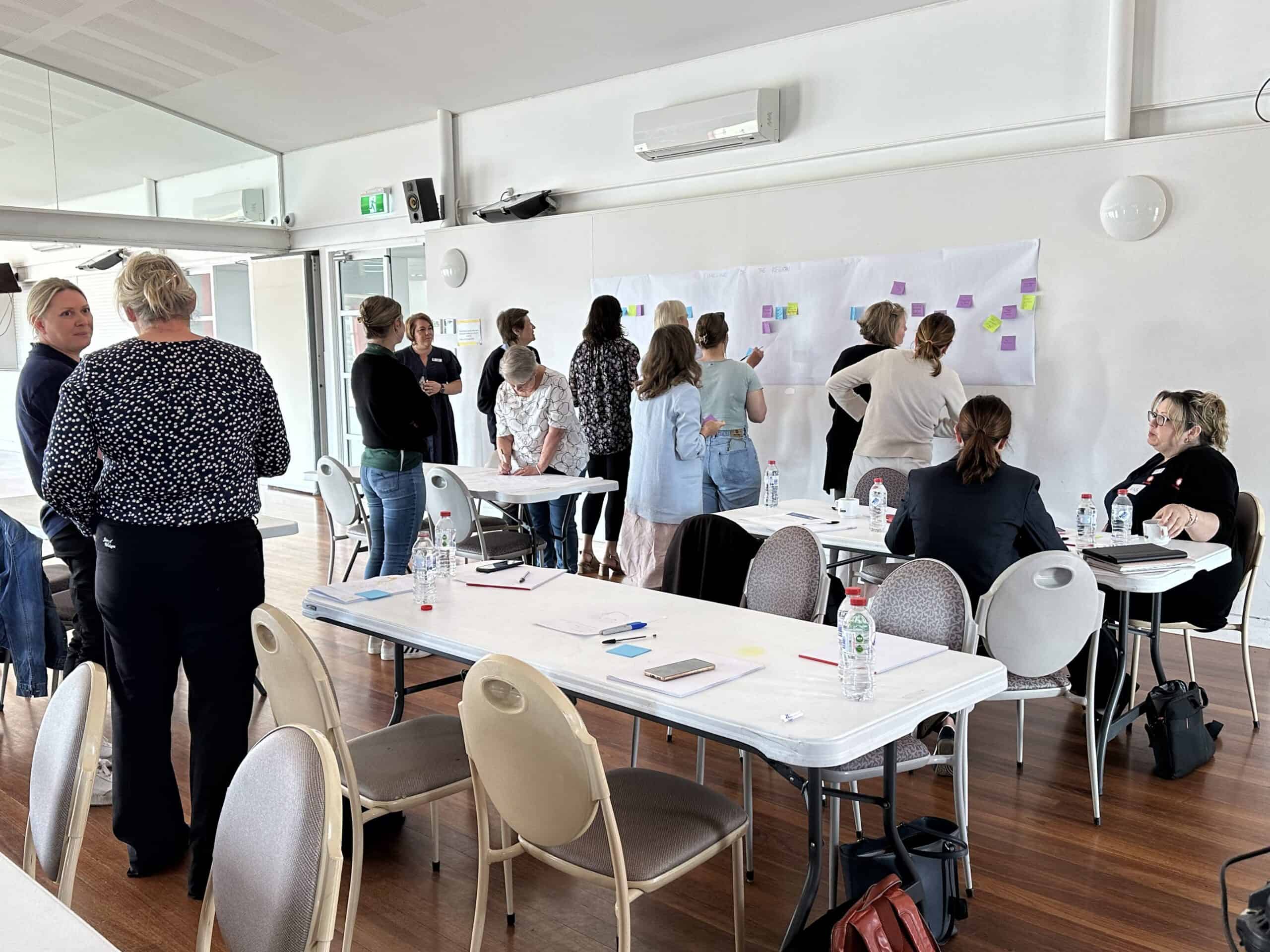 A group of people stand inside a naturally lit room. Their backs are turned to the viewer as they are facing a white board with brainstorming ideas from the Changemaker workshop.