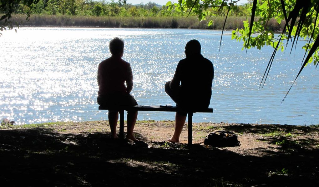 Two men sitting in the shadows in front of a river.